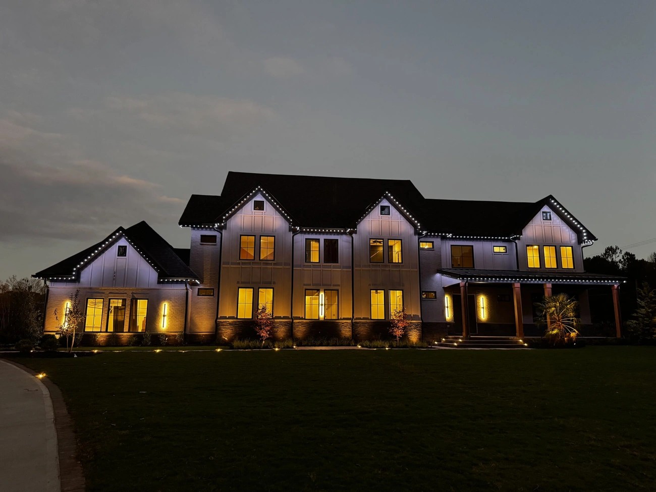 Large modern house at dusk with warm exterior accent lighting highlighting architectural features and landscaping.